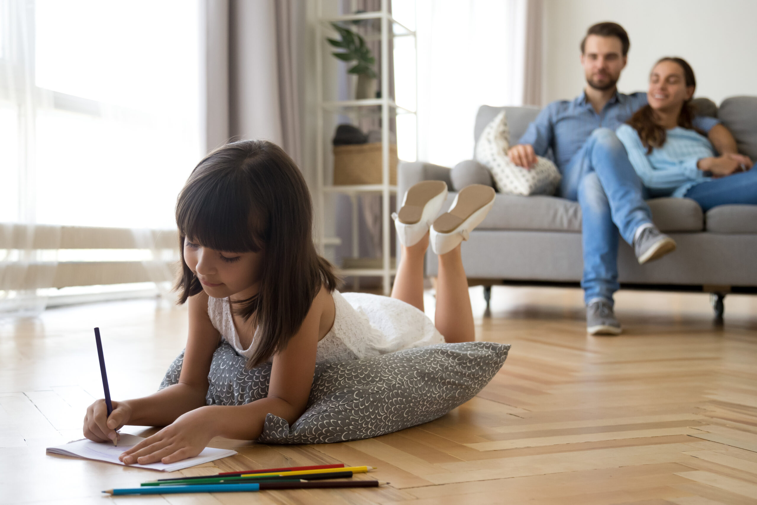 Daughter draws lying on warm floor parents sitting on couch in energy efficient home