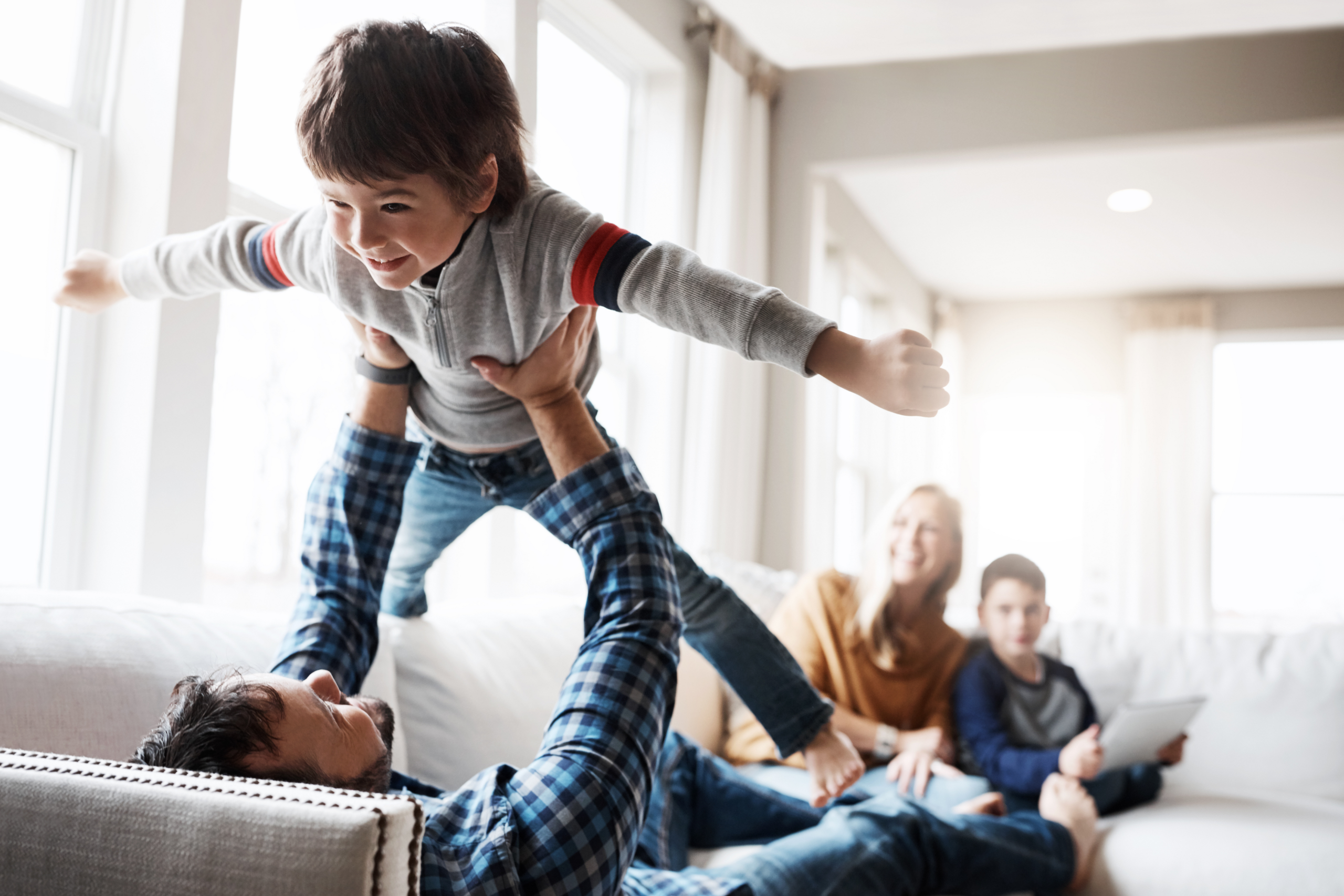 father holding son in the air while lying down on a couch with family watches in background